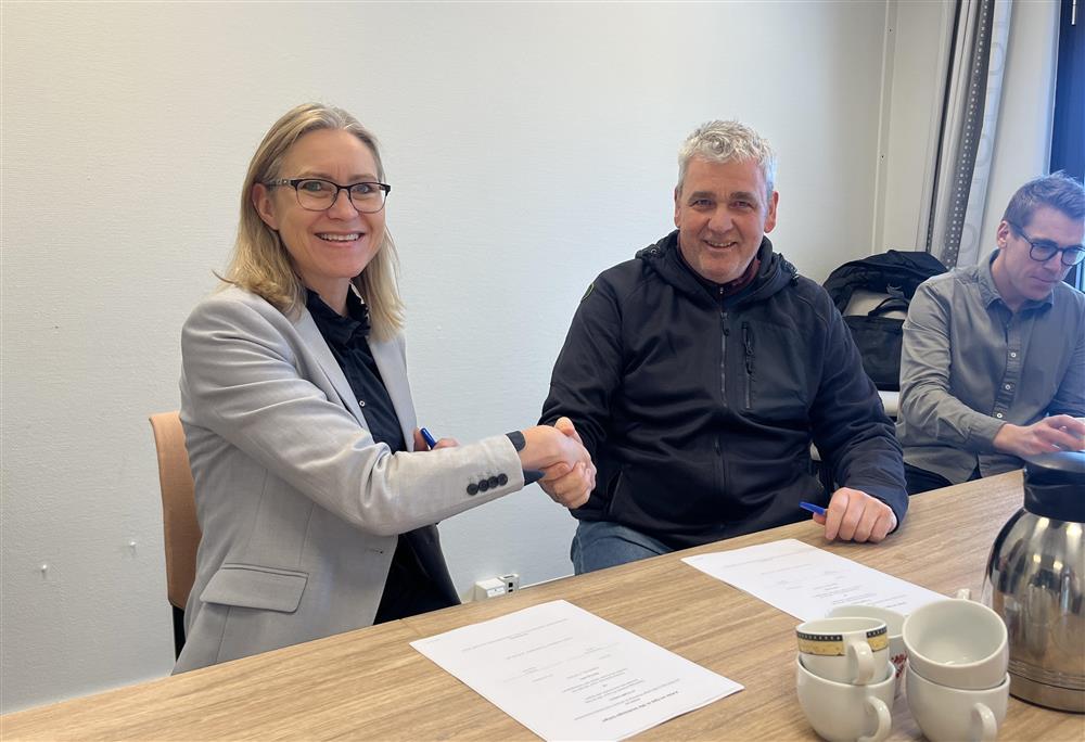 Beate Skretting og Rolf Olsen sitter ved et bord. Kontrakter ligger på bordet og de gir hverandre et håndtrykk.  - Klikk for stort bilde