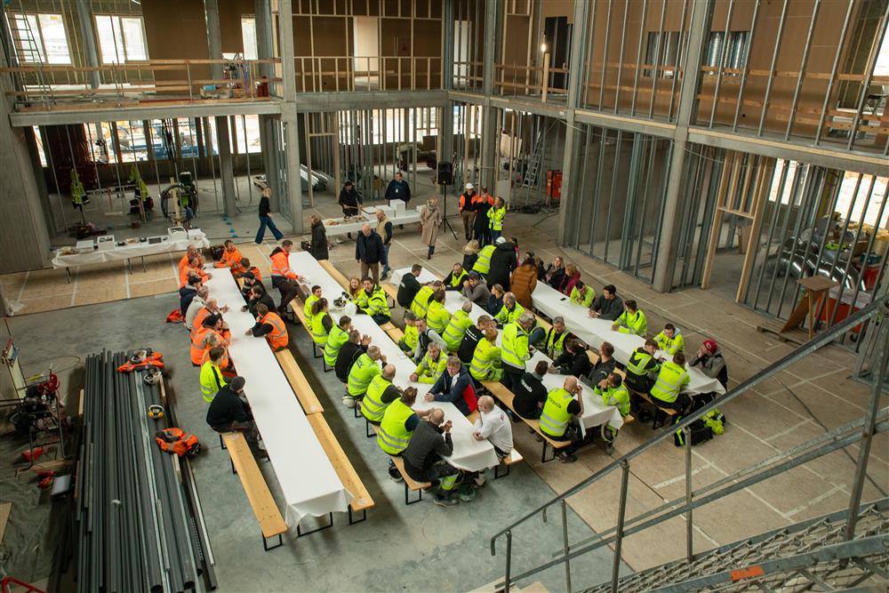 Oversiktsbilde fra 2. etasje på det som skal bli nye Fjære barneskole. Nede på betonggulvet ser vi fire langbord og mange mennesker.  - Klikk for stort bilde