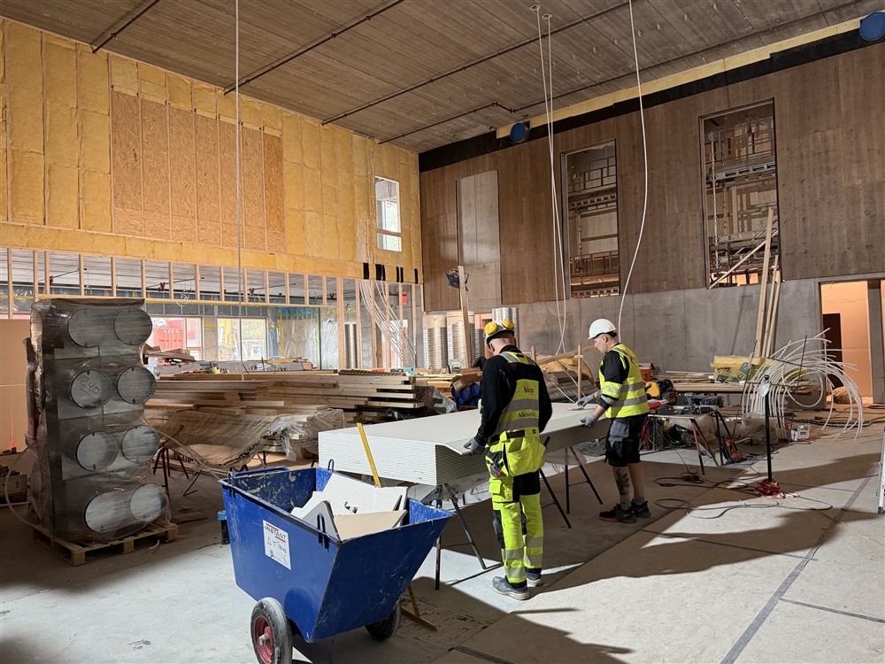 Et stort rom i et bygg under oppføring. Isolerte vegger, stabler med plank, to arbeidere som ser på noen plater.  - Klikk for stort bilde