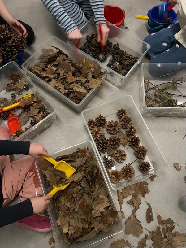Kasser med materialer fra naturen som kongler og blader. Vi ser litt av noen barn som leker med det med spader. - Klikk for stort bilde