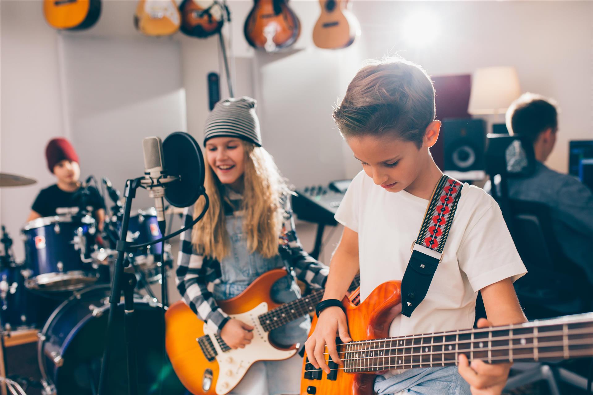 Barn som spiller i band, en på trommer, en på gitar og sang og en på bass. Bak sitter en person ved en datamaskin.