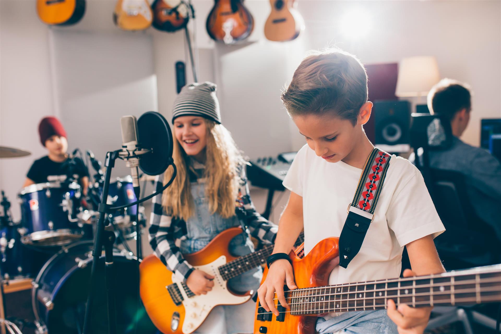 Barn som spiller i band, en på trommer, en på gitar og sang og en på bass. Bak sitter en person ved en datamaskin.