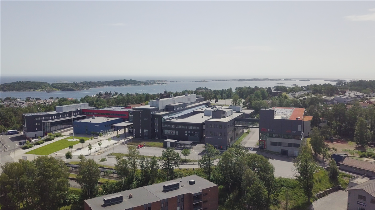 Dronebilde av UiA Campus Grimstad med bygninger, grøntområder og sjøen i bakgrunnen. - Klikk for stort bilde
