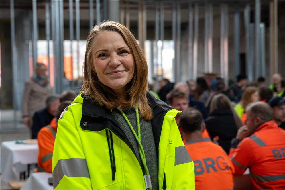 Ida Kristine Steffensen Heiskel smiler. Oppstilt bilde inne i det som skal bli nye Fjære barneskole. Mange arbeidere i bakgrunnen som spiser mat.  - Klikk for stort bilde