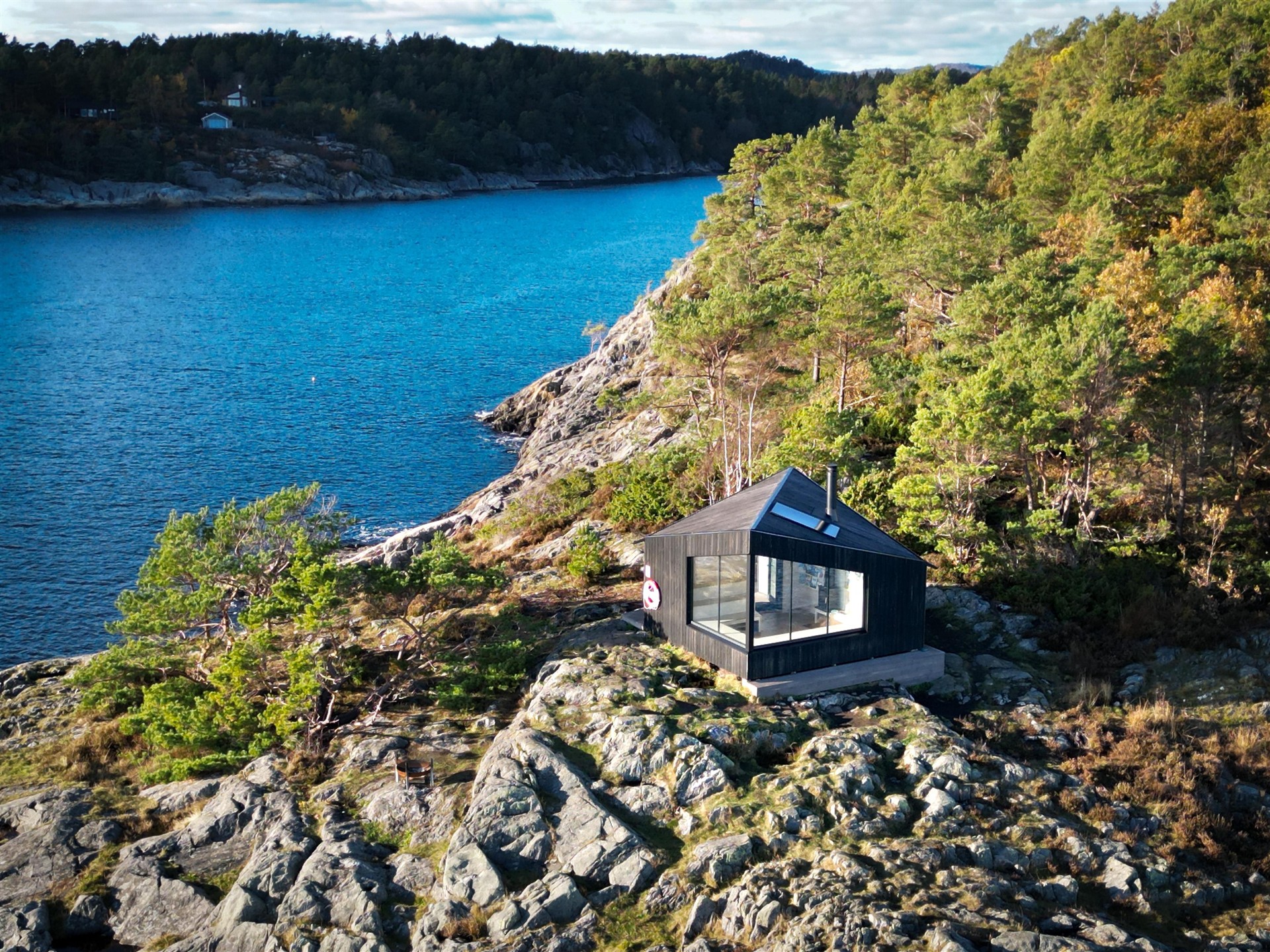 Liten mørk trehytte med store vinduer ligger på svaberg omgitt av furutrær og utsikt mot blått sjøvann i Grimstad.
