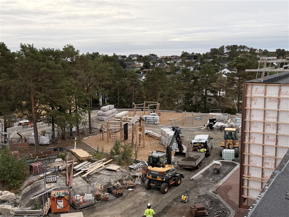 Et lekeområde under oppføring. Lekeplass med lekeapparater, materiell,  trær, biler, gravemaskin og et byggefelt i bakgrunnen.  - Klikk for stort bilde
