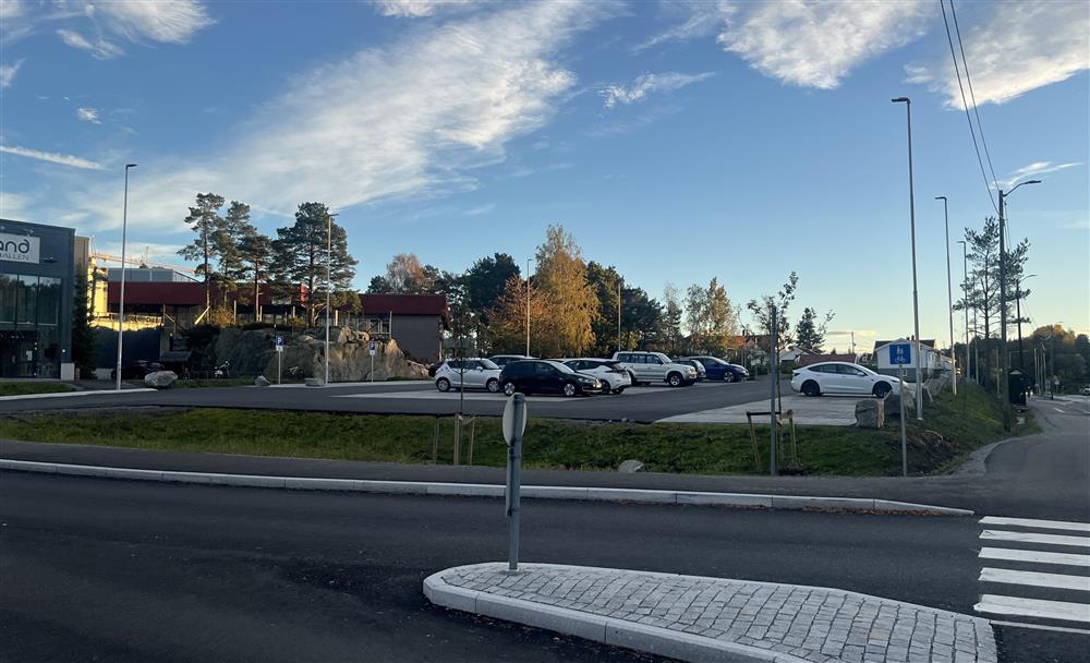 Parkeringsplassen i Holvika med biler, asfalt, trær og turnhallen til venstre - Klikk for stort bilde