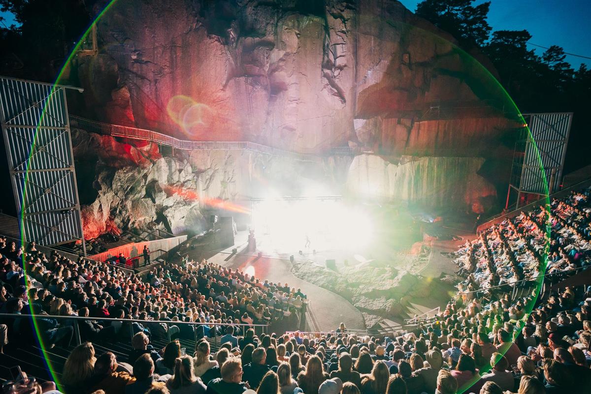 Lyset fra scenen, hvor et band spiller, lyser opp fjellsidene i Fjæreheia Amfi. Fullsatt tribune. - Klikk for stort bilde